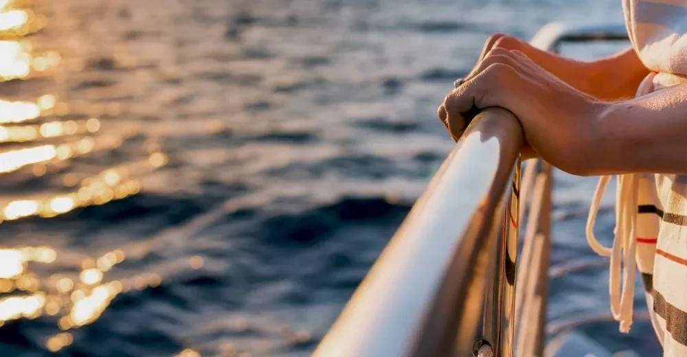 Close up of a person's hands gripping the railing of a boat, silhouetted against a vibrant sunset sky
